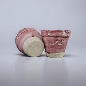 Op de afbeelding: Twee kleine, handgemaakte keramische bloempotten. De potten hebben een stoffige roze kleur met een getextureerde afwerking en een natuurlijke, ongeglazuurde basis. Eén pot staat rechtop en de andere ligt op zijn kant. De tekst "FUCK ME" is in de zijkant van een pot geëtst.