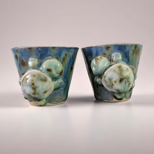 Może przedstawiać: Dwa ręcznie robione ceramiczne kubki z niebieskim szkliwem i teksturowanymi, organicznymi kształtami. Każdy kubek ma skupisko trzech zaokrąglonych, jasno niebieskich i brązowych elementów dekoracyjnych. Kubki mają lekko rozszerzony kształt.