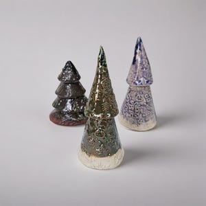 Puede incluir: Tres figuras de cerámica de árboles de Navidad. Una es marrón oscuro con un diseño en capas, otra es verde con motivos florales, y la última es azul con diseños de cajas de regalo y corazones. Los árboles están sobre una superficie blanca.