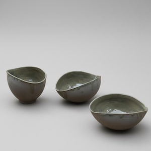 May include: Three handmade ceramic bowls with a unique, organic shape. The bowls are a muted gray-brown color on the exterior, with a light green interior and a white rim. The glaze has a subtle, dripped effect, adding to the handcrafted aesthetic.