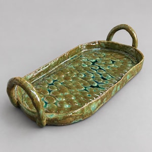 May include: An oval ceramic tray with two arched handles, featuring a glossy glaze in shades of olive green and teal. The surface has a textured, raised pattern of rounded, pebble-like shapes across the base, with a smooth, raised rim edge.