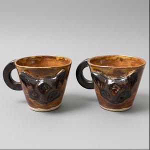 Può includere: Due tazze in ceramica con un disegno a forma di gatto. Le tazze sono marroni con un volto di gatto marrone scuro sul davanti. I manici sono marrone scuro e circolari. Le tazze sono su uno sfondo grigio.