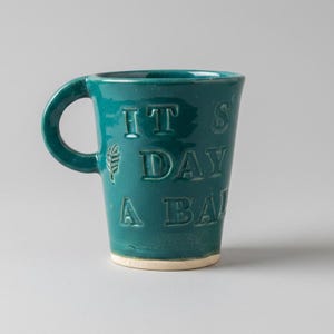 Puede incluir: Una taza de cerámica color verde azulado con acabado brillante y asa redondeada. El frente presenta el texto en relieve IT'S A DAY A BAD, junto con un pequeño icono de hoja en relieve. La base de la taza no está esmaltada y es de color beige.