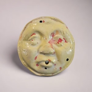 Peut inclure: Un masque en céramique de couleur jaune pâle, représentant un visage aux yeux fermés, un petit nez et une ouverture buccale circulaire. Le masque présente des mouchetures rouges et noires et est probablement fait à la main.