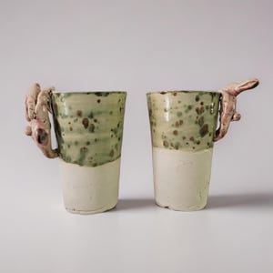 Może przedstawiać: Dwa ceramiczne kubki z unikalnymi uchwytami. Kubki są wysokie i cylindryczne, z dwutonowym wzorem. Dolna połowa jest kremowobiała, a górna połowa jest nakrapiana na zielono. Jeden kubek ma uchwyt w kształcie dłoni, a drugi w kształcie królika.
