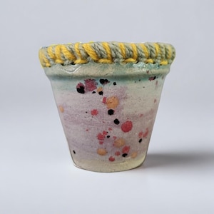 May include: A small, hand-painted ceramic pot with a speckled design in shades of pink, orange, and black. The rim is adorned with a yellow and grey woven trim. The pot has a light blue and cream base.