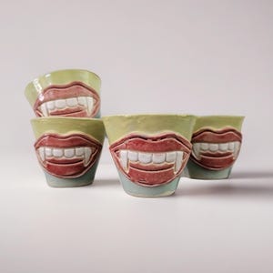 May include: Four ceramic cups with a unique design. Each cup features a light green rim, a red mouth with white teeth, and a light blue base. The cups are arranged on a white surface, showcasing their distinctive vampire-inspired aesthetic.