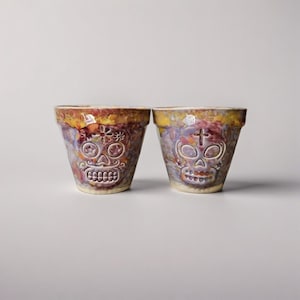 Könnte beinhalten: Zwei Keramik-Blumentöpfe mit einem Totenkopf-Design zum Tag der Toten. Die Töpfe haben eine strukturierte, glasierte Oberfläche in Lila, Gelb und Orange. Einer hat ein Blumendesign, der andere ein Kreuz.