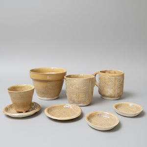 Könnte beinhalten: Eine Sammlung handgefertigter, beigefarbener Keramik-Tischutensilien. Das Set umfasst einen großen Pflanztopf, einen Becher mit Henkel, einen kleineren Becher, Untertassen und kleine Teller. Jedes Stück hat ein Reliefdesign mit Libellen und Blumen.