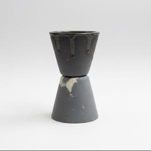 Puede incluir: Un recipiente cerámico apilado con un acabado gris oscuro texturizado. La pieza superior es una pequeña taza acampanada con gotas oscuras. La pieza inferior es una base en forma de cono. Sobre un fondo blanco.