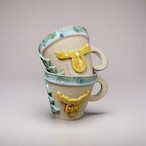 May include: Two stacked ceramic mugs. The mugs are off-white with a light blue rim and a yellow, abstract design. One mug has a handle. The mugs are handmade and have a rustic, artistic appearance.