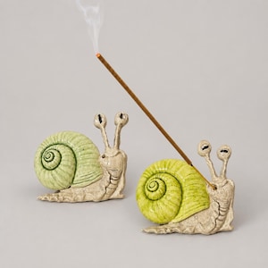Peut inclure: Deux porte-encens en céramique en forme d'escargot avec des coquilles vert clair et des corps beiges. Un escargot porte un bâton d'encens allumé inséré dans sa coquille, émettant un fin filet de fumée blanche. Les deux ont des coquilles en spirale.