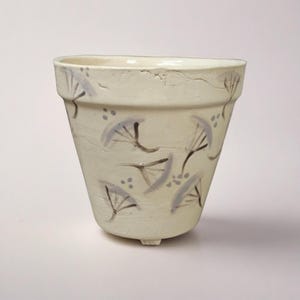 May include: Cream-colored ceramic planter with a slightly textured surface. The planter features a hand-painted design of stylized, gray-blue dandelion seed heads. The planter has a wide top, a tapered body, and three small feet.