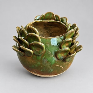 Op de afbeelding: Een ronde keramische pot met een glanzend groen glazuur, voorzien van talloze kleine, overlappende, bloembladachtige klei-uitsteeksels op het buitenoppervlak. De pot heeft een open bovenkant en een gladde, afgeronde basis met bruine tinten.