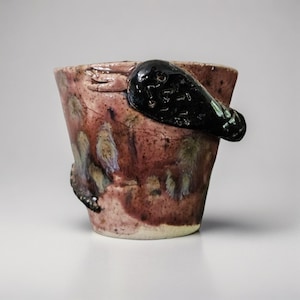 May include: A handcrafted ceramic bucket with a dark handle and a textured, mottled surface. The bucket has a reddish-brown base colour with streaks of yellow, purple, and black. A small hand is sculpted on the side.