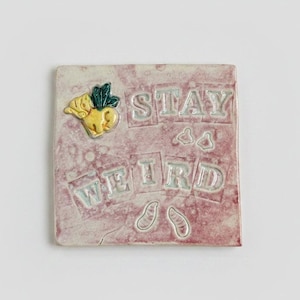 May include: A square ceramic tile with a mottled pink and white glaze. The text STAY WEIRD is embossed in block letters. A small yellow creature with green wings is positioned in the top left corner, accompanied by small embossed shapes resembling footprints.