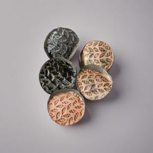 May include: A collection of five ceramic coasters. Two are black with a textured leaf pattern, and three are tan with a similar leaf design. The coasters are round and have a rustic, handmade appearance.
