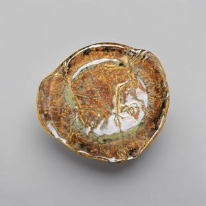 Pode incluir: Um prato de cerâmica em forma de ostra com uma borda dourada. O interior apresenta um padrão salpicado de tons castanhos, dourados e verdes. O prato tem um acabamento brilhante e está sobre um fundo cinzento.