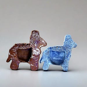 May include: Two ceramic donkey-shaped planters. One is brown with purple accents, the other is blue. Both have a detailed floral pattern and a central pocket for plants. The planters are decorative and suitable for small plants.