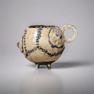 May include: A handcrafted ceramic mug with a unique, textured design. The beige mug features black stitching details and a circular handle. It stands on three small legs, adding to its artistic appeal. The mug's surface has a rough, tactile quality.