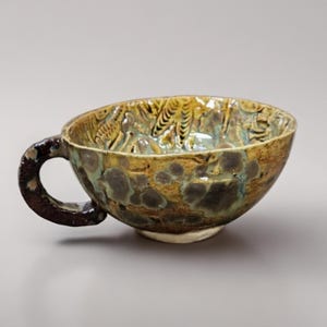 Peut inclure: Tasse à thé en céramique faite à la main avec une anse marron foncé et un glaçage texturé et multicolore. La tasse présente un mélange de teintes marron, jaune et verte, avec un motif détaillé à l'intérieur. La base est d'une couleur plus claire, non émaillée.