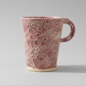 May include: A ceramic mug with a tapered cylindrical body and a rounded handle. The exterior features a textured, embossed floral pattern in shades of dusty rose and cream, with a smooth, unglazed base rim and a deep pink interior finish.