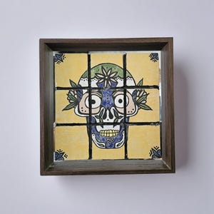 May include: A framed artwork featuring a Day of the Dead skull design. The skull is painted on nine yellow tiles with blue, green, and white details. The artwork is encased in a dark brown wooden frame, with decorative corner accents.