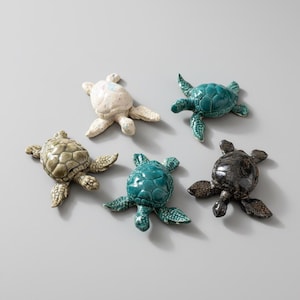 Könnte beinhalten: Fünf Keramik-Schildkrötenfiguren in verschiedenen Farben: Creme, Türkis, Olivgrün, Dunkeltürkis und Schwarz mit Goldglitter. Jede Schildkröte hat einen strukturierten Panzer und flossenartige Beine.