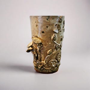 May include: A tall, beige ceramic mug with a textured surface and a detailed relief design. The design includes mushrooms, leaves, and flowers. The mug has a rustic, handmade appearance, suggesting it is a piece of pottery.