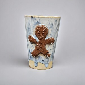 May include: A handmade ceramic tumbler with a brown gingerbread man design. The tumbler has a white and blue glaze with a dripped effect. The base of the tumbler is a light beige color. The gingerbread man has an angry expression.