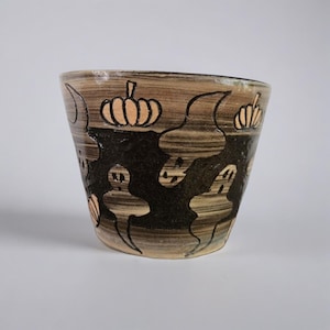 May include: A ceramic Halloween-themed bowl with a brown and black design. The bowl features hand-drawn pumpkins and ghost-like figures. The bowl has a slightly irregular shape and a light-colored interior.