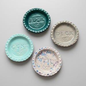 Puede incluir: Cuatro platos decorativos de cerámica con bordes festoneados. El plato verde oscuro presenta la palabra FUCK, el azul claro CUNT, el beige DICK y el blanco moteado BITCH, cada uno con un pequeño corazón grabado en la base.