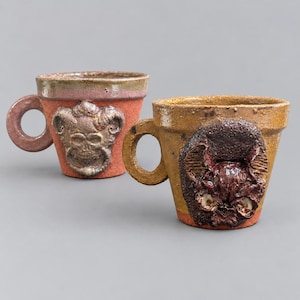 May include: Two ceramic mugs with circular handles. The left mug is reddish-brown with a sculpted skull emblem. The right mug is mustard-yellow with a dark, textured bat-like creature relief. Both feature a tapered shape and glazed finish.