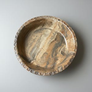 Könnte beinhalten: Eine runde, dekorative Schale mit einem marmorierten Muster in Braun-, Beige- und Grautönen. Der Rand weist ein sich wiederholendes Blumendesign auf. Das Innere der Schale hat eine glänzende Oberfläche, und die Gesamtästhetik ist rustikal und erdig.