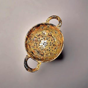 May include: A gold-toned colander with two loop handles. The bowl has a floral pattern and small drainage holes. The colander has a glossy finish. The item is set against a neutral background.