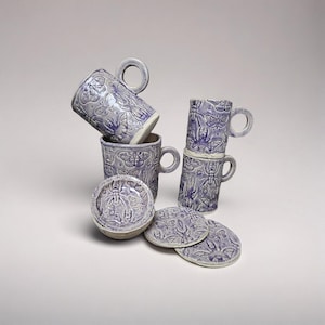 Può includere: Un set di tazze e sottobicchieri in ceramica lavanda. Le tazze, con manico, sono decorate con disegni a rilievo di insetti e farfalle. I sottobicchieri sono rotondi e presentano anche disegni di insetti e farfalle.