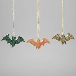 Peut inclure: Trois ornements de chauve-souris en céramique, verts, oranges et dorés, suspendus à des ficelles. Les chauves-souris présentent des motifs d'ailes détaillés, parfaits pour Halloween. Chaque chauve-souris a une couleur et un design uniques.