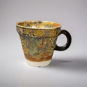 Puede incluir: Una taza de cerámica hecha a mano con un diseño texturizado único. La taza presenta un asa marrón y un cuerpo con una mezcla de colores marrón, verde y beige. La base es blanca y el borde es marrón oscuro.