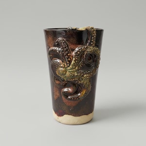 Puede incluir: Un vaso alto de cerámica con un diseño detallado de pulpo. La taza marrón oscuro presenta un pulpo texturizado y en relieve con detalles verdes y dorados. La base es de color beige claro.
