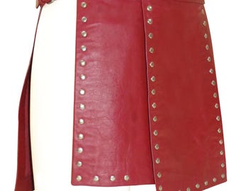 Op de afbeelding: Een rode leren rok met zilverkleurige klinknagels langs de randen. De rok heeft een riem met een zilveren gesp. Het ontwerp heeft een overlappend voorpaneel.