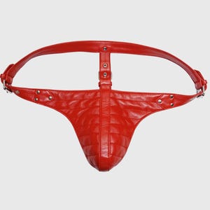 Puede incluir: Arnés de cuero rojo con diseño acolchado. El arnés presenta una correa que rodea la cintura y una correa vertical con detalles de hebilla. El diseño incluye remaches metálicos y una forma curva.