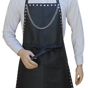 Puede incluir: Delantal de cuero negro con tachuelas plateadas y detalle de cadena. El delantal se ata a la cintura y se lleva sobre una camisa blanca. Adecuado para diversas actividades.