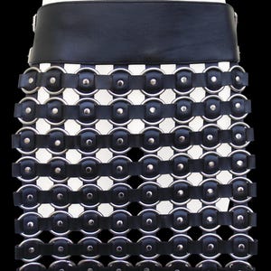 Puede incluir: Falda negra y plateada con una ancha cinturilla de cuero negro. La falda presenta un patrón de rejilla de anillos plateados conectados por correas de cuero negro, creando un diseño único. La falda está sobre un fondo negro.