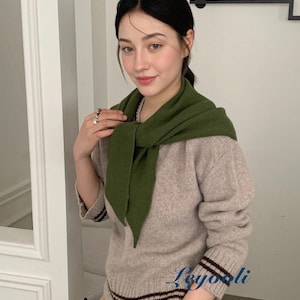 May include: A woman wearing a beige sweater with brown stripes at the cuffs and hem, paired with khaki pants. A green scarf is draped around the neck. The image is taken indoors.