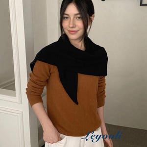 May include: A woman wearing a black scarf over a brown sweater and white trousers. The sweater has long sleeves. The woman is smiling and holding a beige bag. The background is a neutral colour.
