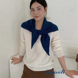 May include: A woman wearing a cream-colored sweater with a navy blue scarf draped around her neck. She is also wearing brown pants. The sweater has long sleeves and a crew neck. The scarf is tied in a knot at the front.