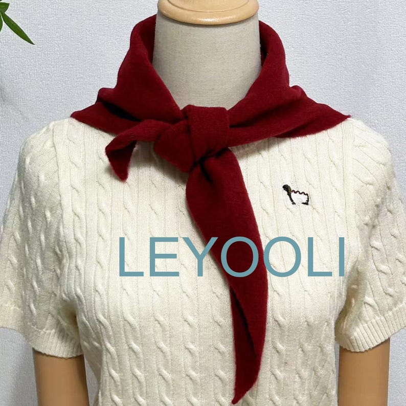 May include: A red knitted scarf tied in a knot around the neck of a white cable knit sweater. The scarf is tied in a loose knot and hangs down the front of the sweater. The sweater has a small embroidered sheep on the front. The text "LEYOOLI" is printed on the sweater.