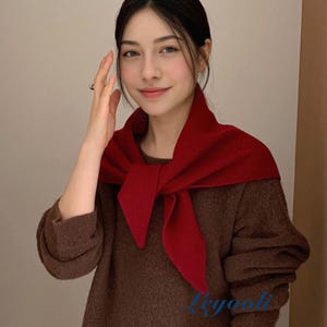 May include: A woman wearing a dark red scarf over a brown sweater. The scarf is knotted at the front, creating a layered look. The sweater has long sleeves and a crew neck. The background is a neutral beige colour.