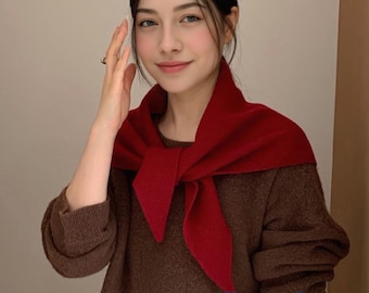 May include: A woman wearing a dark red scarf over a brown sweater. The scarf is tied in a knot at the front, creating a layered look. The sweater has long sleeves and a crew neck. The background is a neutral beige color.