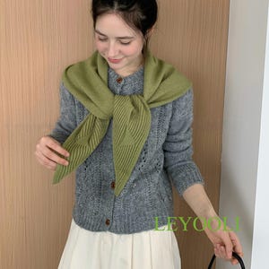以下が含まれることがあります： 木製のボタンが付いたグレーのカーディガンとグリーンのスカーフ。白いパンツと合わせています。「LEYOOLI」の文字が緑色で見えます。